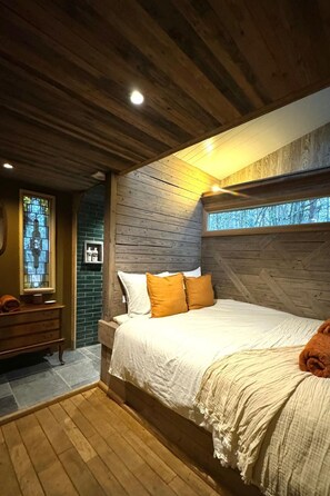 A bedroom with a wooden ceiling, a bed with white bedding and orange pillows, a dresser, and a window with a view of trees. - Camping Ont Moeten - Rumi by Interhome (Rekken)