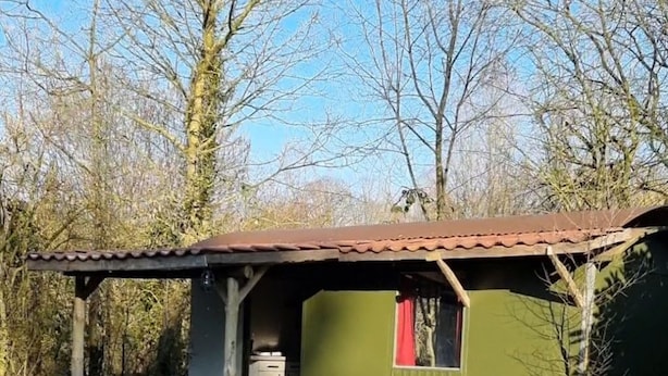 Ein kleines, eingeschossiges Gebäude mit grüner Fassade und braunem Dach. Es besitzt eine kleine Veranda mit Tisch und Stühlen sowie ein Fenster mit rotem Vorhang.