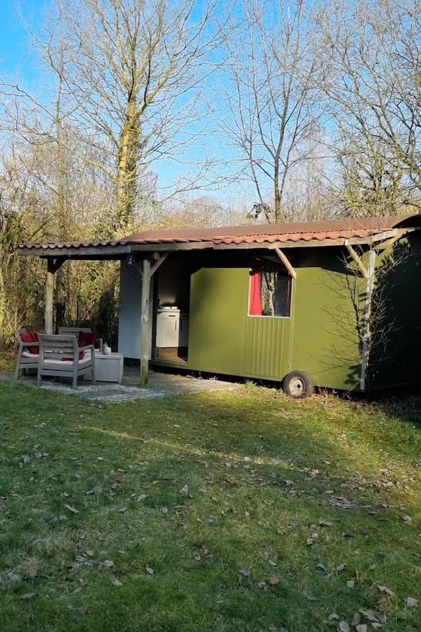 A small, single-story building with a green exterior and a brown roof. It features a small porch with a table and chairs, and a window with a red curtain. - Camping Ont Moeten - T piept zo by Interhome (Rekken)