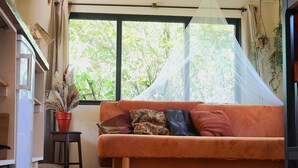 Ein Zimmer mit einer Couch, einem kleinen Tisch, einer Topfpflanze und einem Fenster mit Blick auf Bäume.