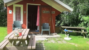 Une petite construction en bois avec un banc et une table de pique-nique, une porte avec une pancarte et une chaise à l’extérieur.