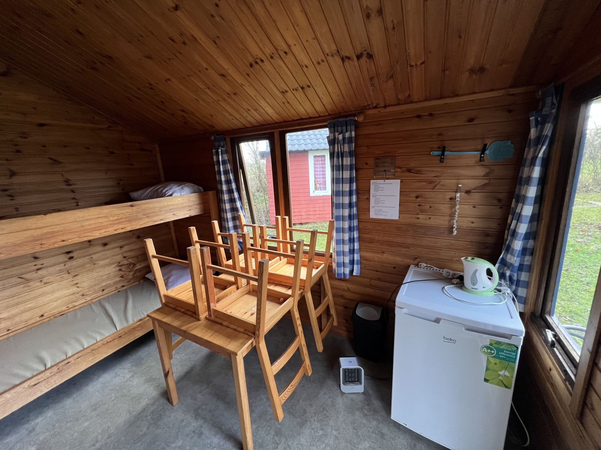 Interior de una cabaña de madera con literas, una mesa de comedor, sillas, un refrigerador y una ventana con cortinas.