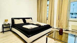 A modern bedroom with a black and white bed, a glass coffee table, and a view of buildings through large windows. - 3BR for 9 Near FAC Platz (Vienna)