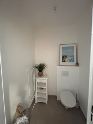 Ein kleines, weißes Badezimmer mit Toilette, einem gerahmten Bild an der Wand, einem kleinen Regal mit einer Pflanze und einem Handtuchhalter.