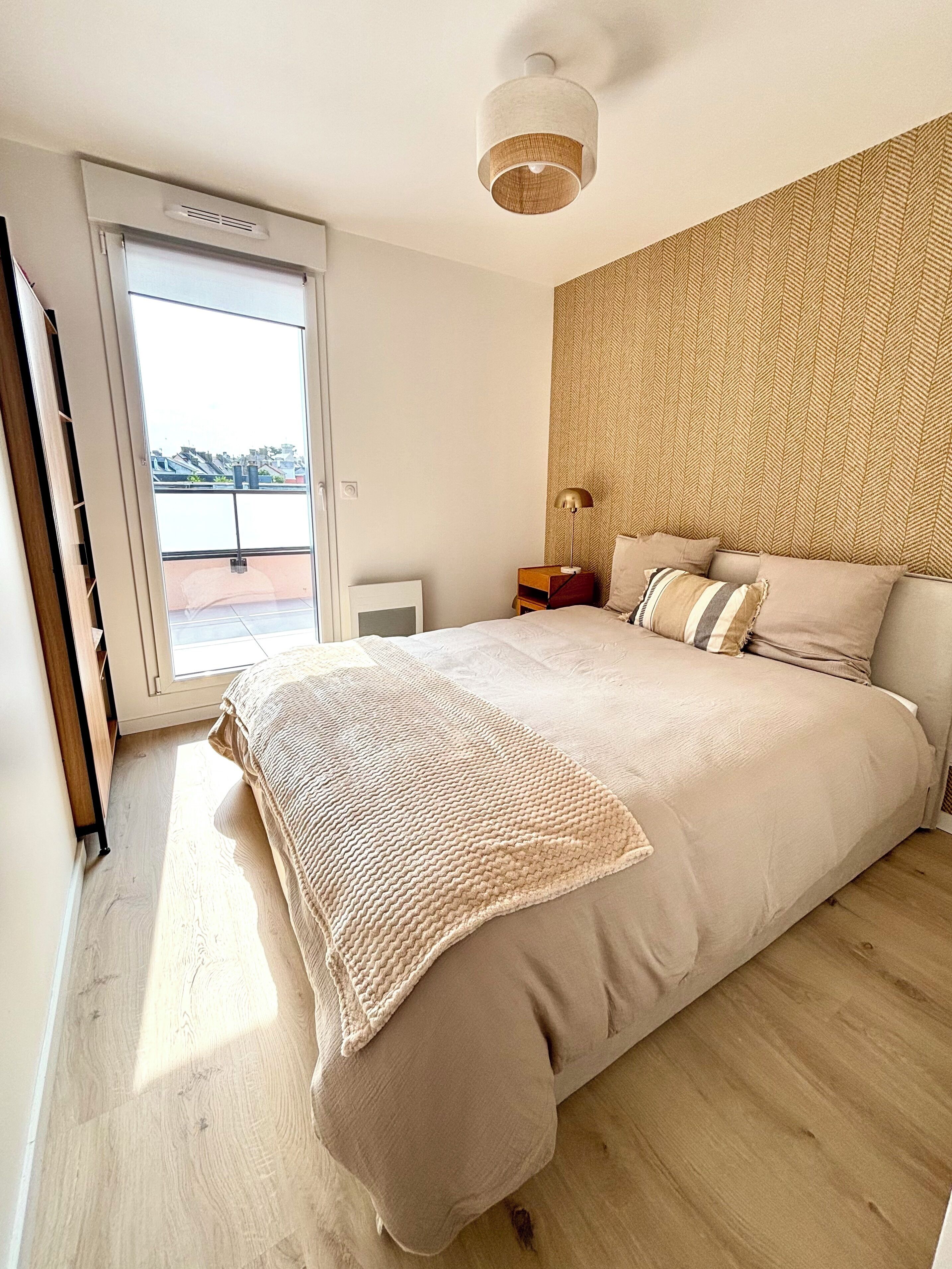 A bedroom with a large bed, a wooden headboard, a bedside table with a lamp, and a view through a sliding door to the outside.