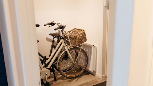 A bicycle with a basket, parked in a hallway with a radiator and a wooden floor.