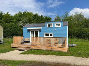 A small, single-story blue house with a deck, a porch, and a gravel path leading to the entrance. - De Glamping - Blauwe Tiny House | 6p by Interhome (Zeewolde)