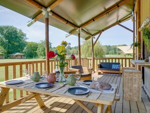 An outdoor wooden deck with a table set for dining, a sofa, and a grill. - De Glamping - Sunny Lodge | 6p by Interhome (Zeewolde)