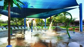 A water play area with multiple water jets, a covered seating area, and a surrounding landscape with trees and a fence.