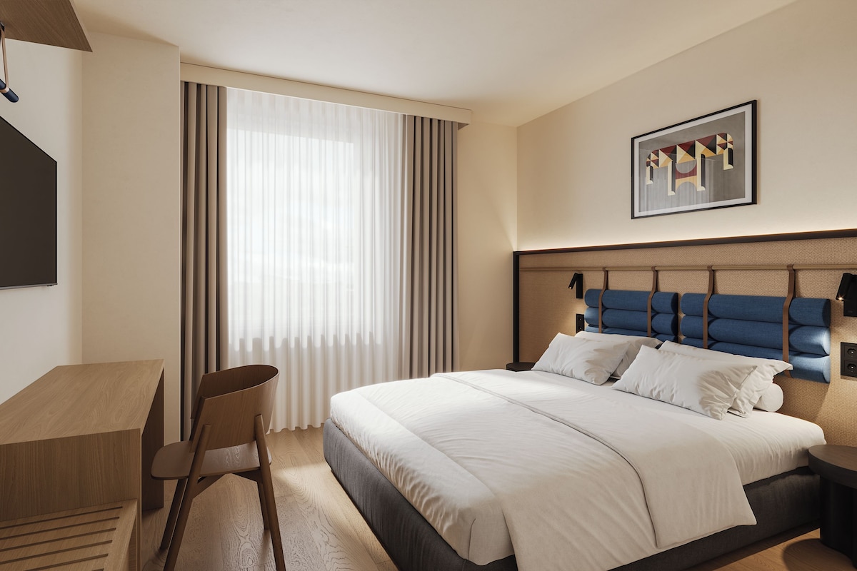 Habitación de hotel con cama, escritorio, silla, televisor montado en la pared y ventana con cortinas.