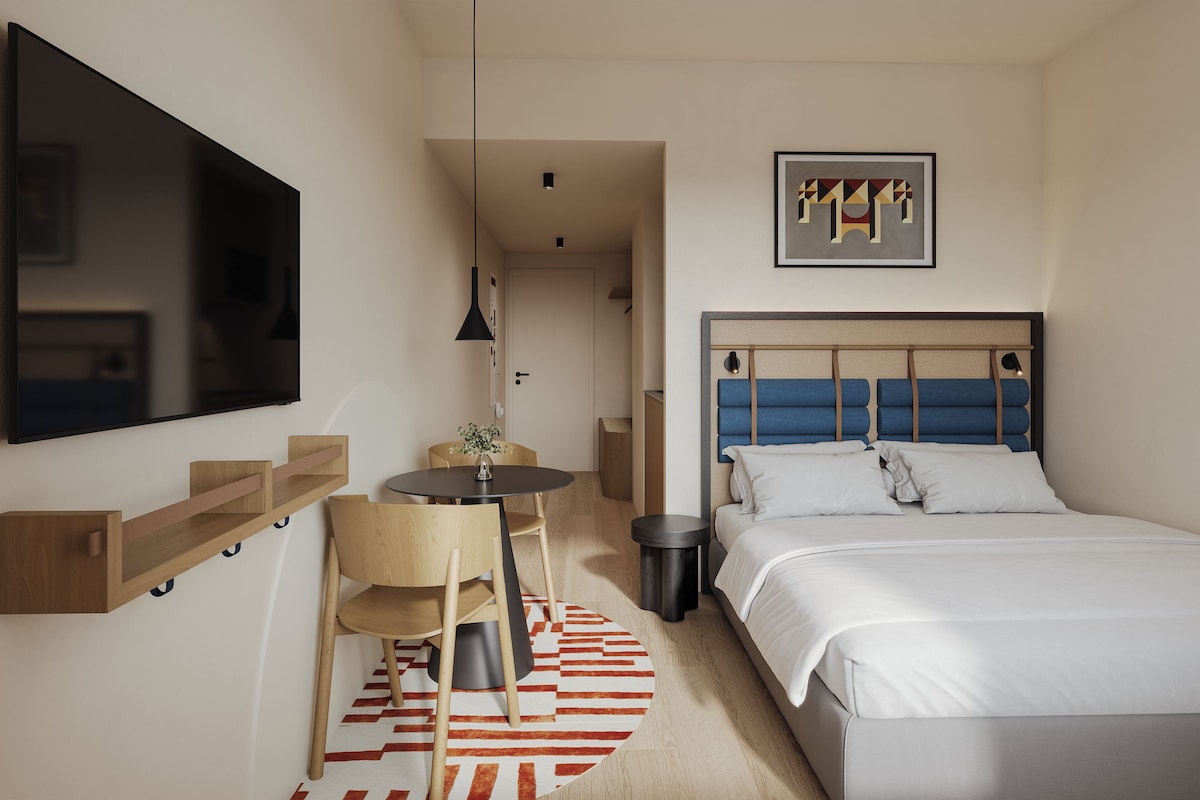 A hotel room with a bed, a desk, a chair, a TV, and a framed artwork on the wall.