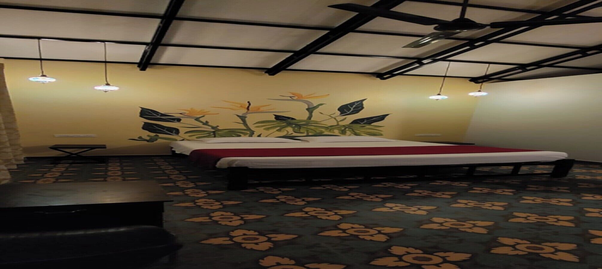 Een hotelkamer met een bed, een tafeltje en een bankje. Er is een plafondventilator en hanglampen.
