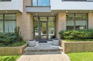A modern building entrance with a glass door, a brick facade, and a small garden area with planters.
