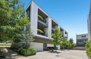 A multi-story residential building with balconies, surrounded by greenery and a clear sky.