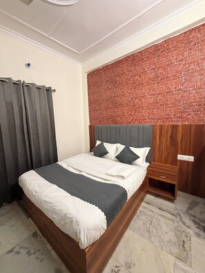 A neatly made bed with a grey and white duvet, a wooden nightstand with a drawer, and a brick-patterned wall behind the bed. - Janki Homestay Rishikesh Laxman Jhula (Rishikesh)