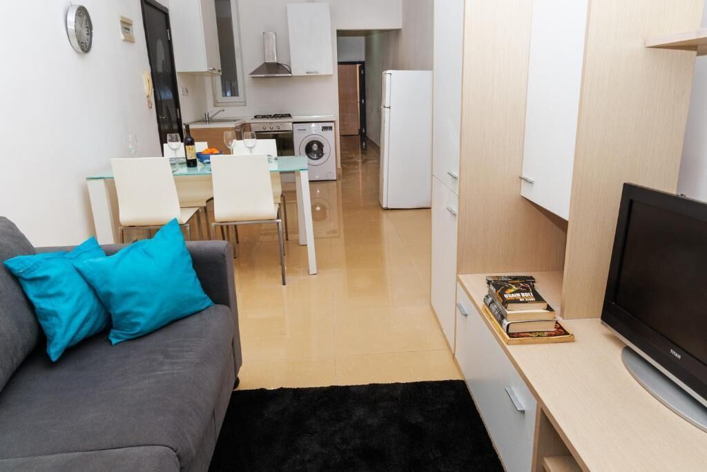 A compact living space with a sofa, a flat-screen TV, a dining area with chairs, a kitchen with appliances, and a washing machine.