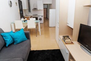 Un espacio habitable compacto con sofá, televisor de pantalla plana, zona de comedor con sillas, cocina equipada y lavadora.
