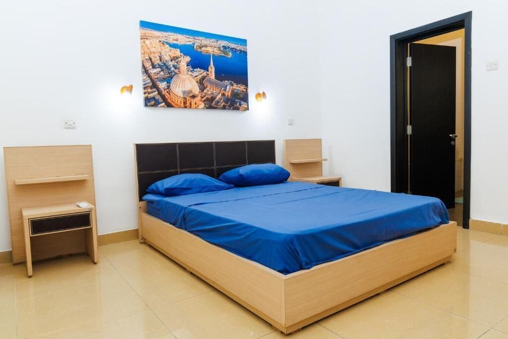 A bedroom with a bed, a nightstand, a wall-mounted lamp, and a door.