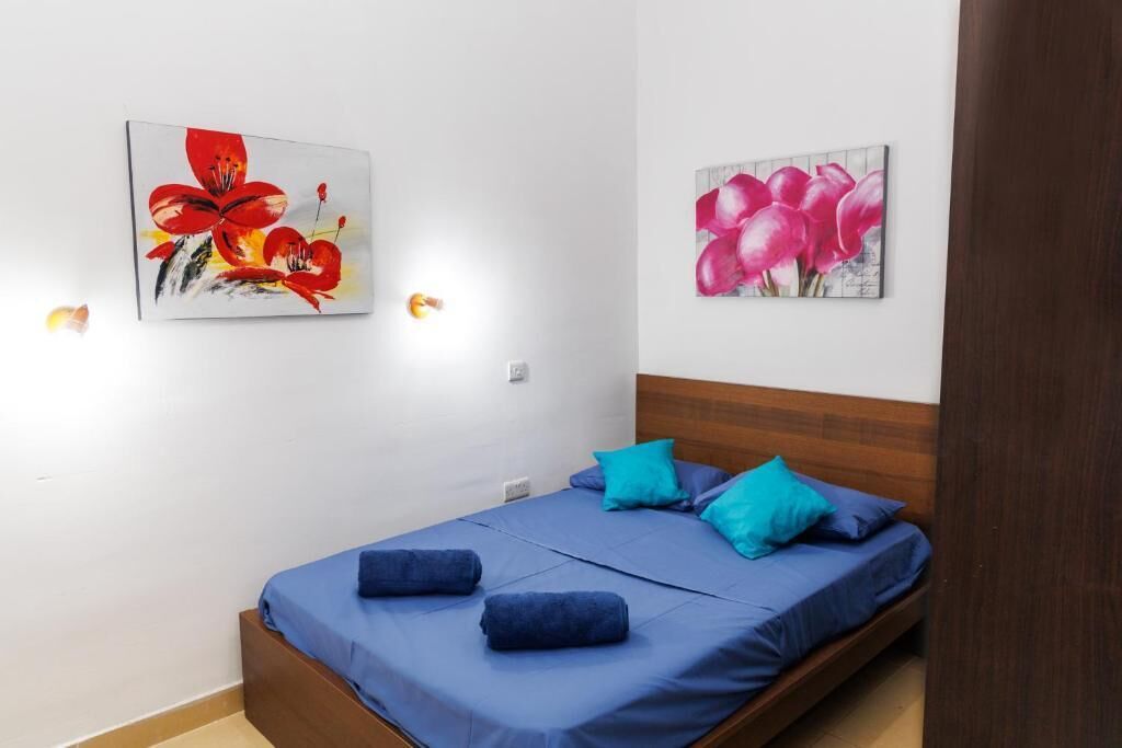 A single bed with blue bedding and pillows, a wooden headboard, and two wall-mounted paintings of flowers.