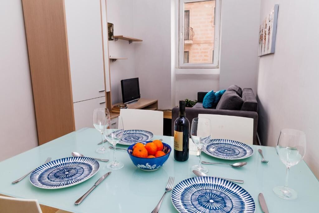 A dining area with a table set for four, a bowl of oranges, a bottle of wine, and a television in the background.