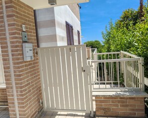 Une entrée fermée par un portail blanc, un mur de briques et un bâtiment avec des volets.