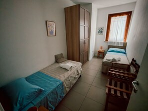 Une petite pièce, de taille modeste, comprenant deux lits simples, une armoire en bois, une petite table avec une lampe et une fenêtre avec des rideaux.