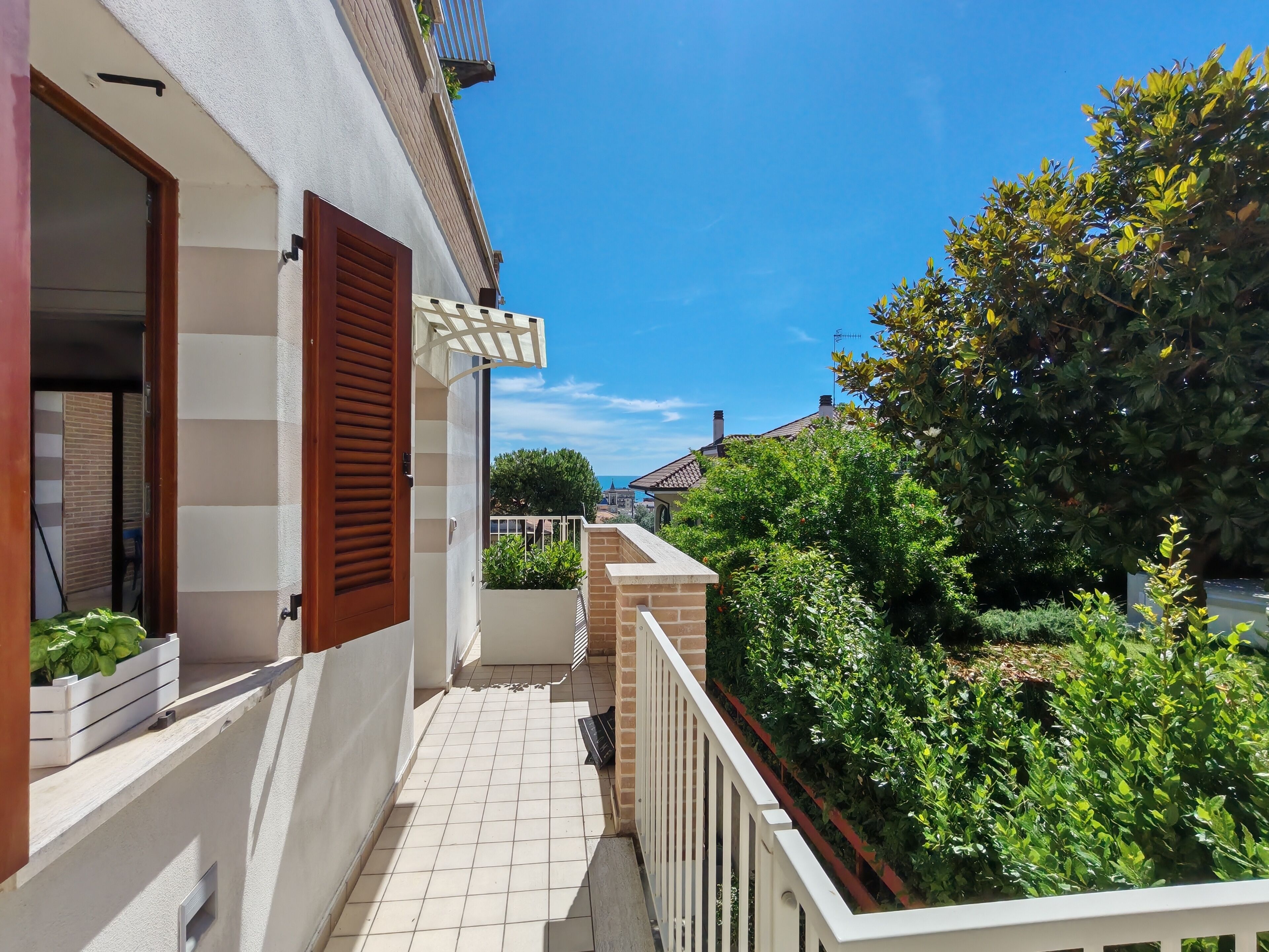 Ein Wohngartenbalkon mit weißer Brüstung, einer Holztür und Blick auf einen Garten sowie Nachbarhäuser.