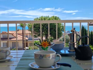 Un balcon avec une table préparée pour le thé, une vue sur la mer et des bâtiments en arrière-plan.