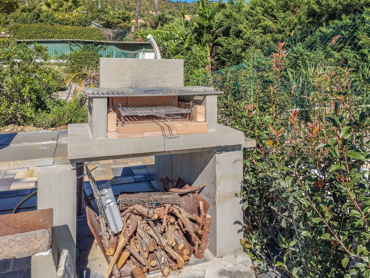 An outdoor grill with a concrete structure, surrounded by greenery.