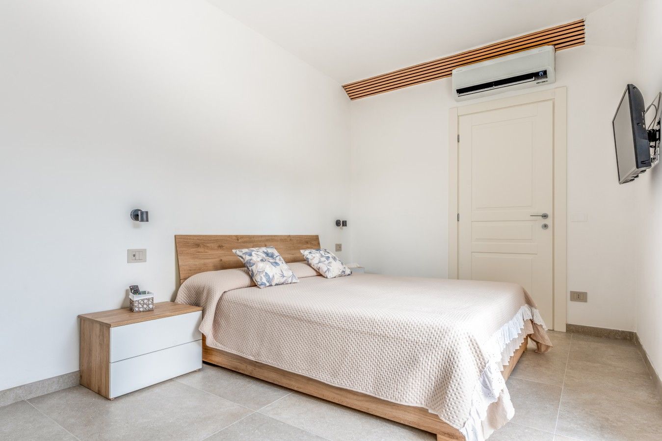 A bedroom with a wooden bed frame, a bedside table, a nightstand, a wall-mounted air conditioner, and a door.