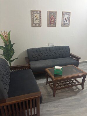 A living room with a dark-colored sofa, a glass coffee table with a green object on it, and two framed pictures on the wall. - Beautiful 3-BHK  with Balcony in Sahibzada Ajit Singh Nagar Chandigarh Airport (Sahibzada Ajit Singh Nagar)