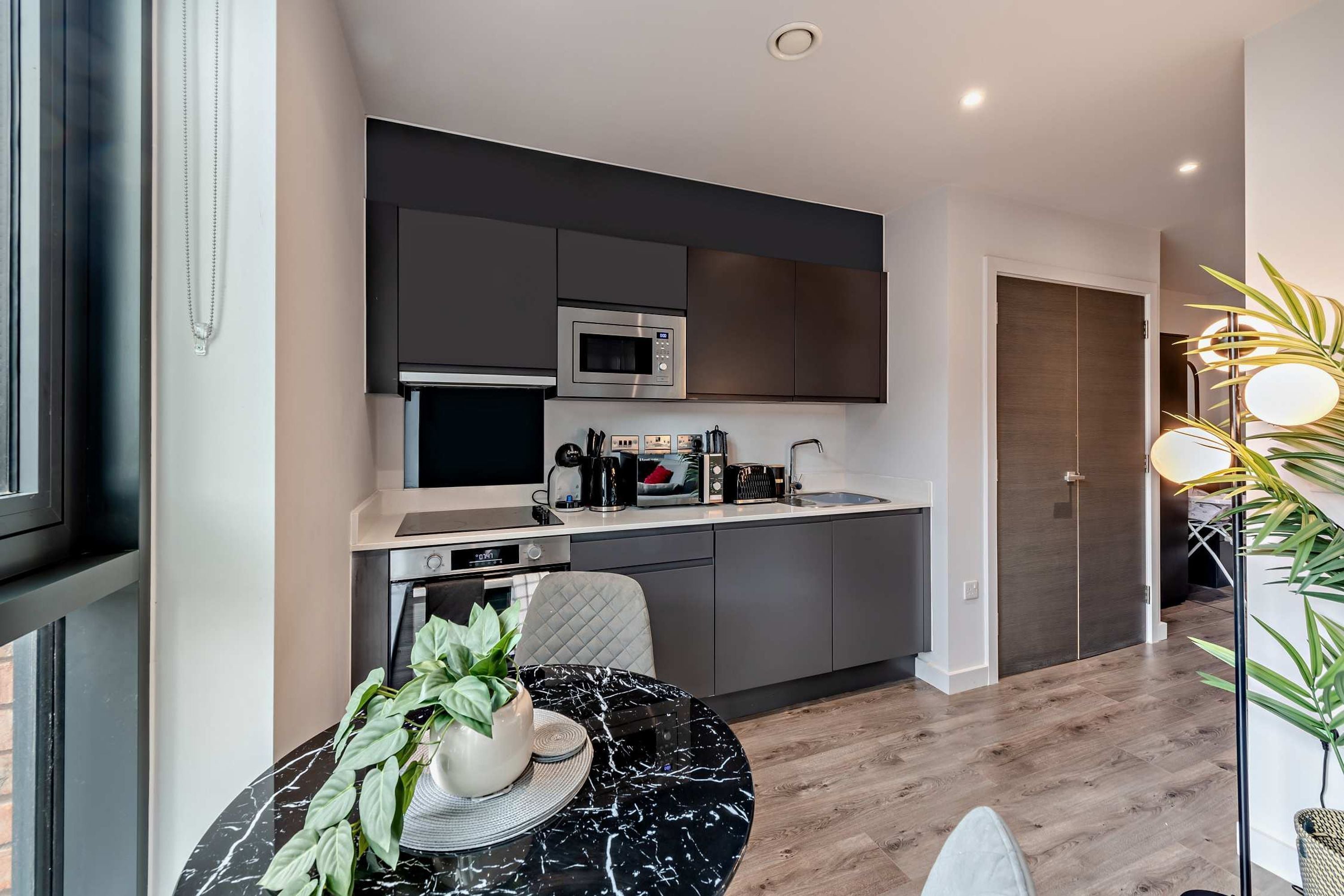 A modern kitchen with a microwave, oven, and coffee maker. There is a dining table with a plant and a chair. The kitchen has dark cabinets and a white countertop.