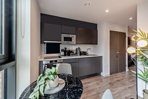 A modern kitchen with a microwave, oven, and coffee maker. There is a dining table with a plant and a chair. The kitchen has dark cabinets and a white countertop. - Host Stay Miller s Place Apartment (Liverpool)