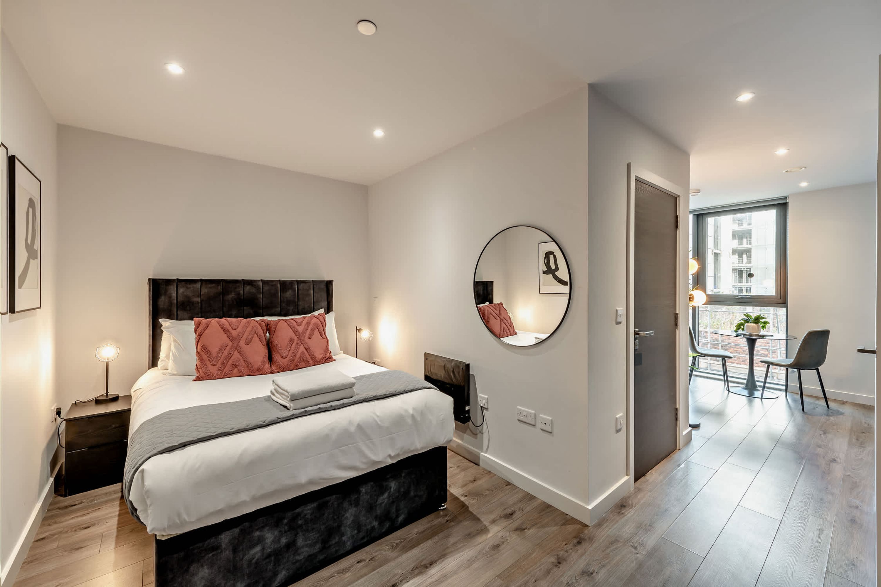 Quarto moderno com cama grande, luminárias laterais, espelho redondo e vista para uma sala de jantar com mesa e cadeiras.