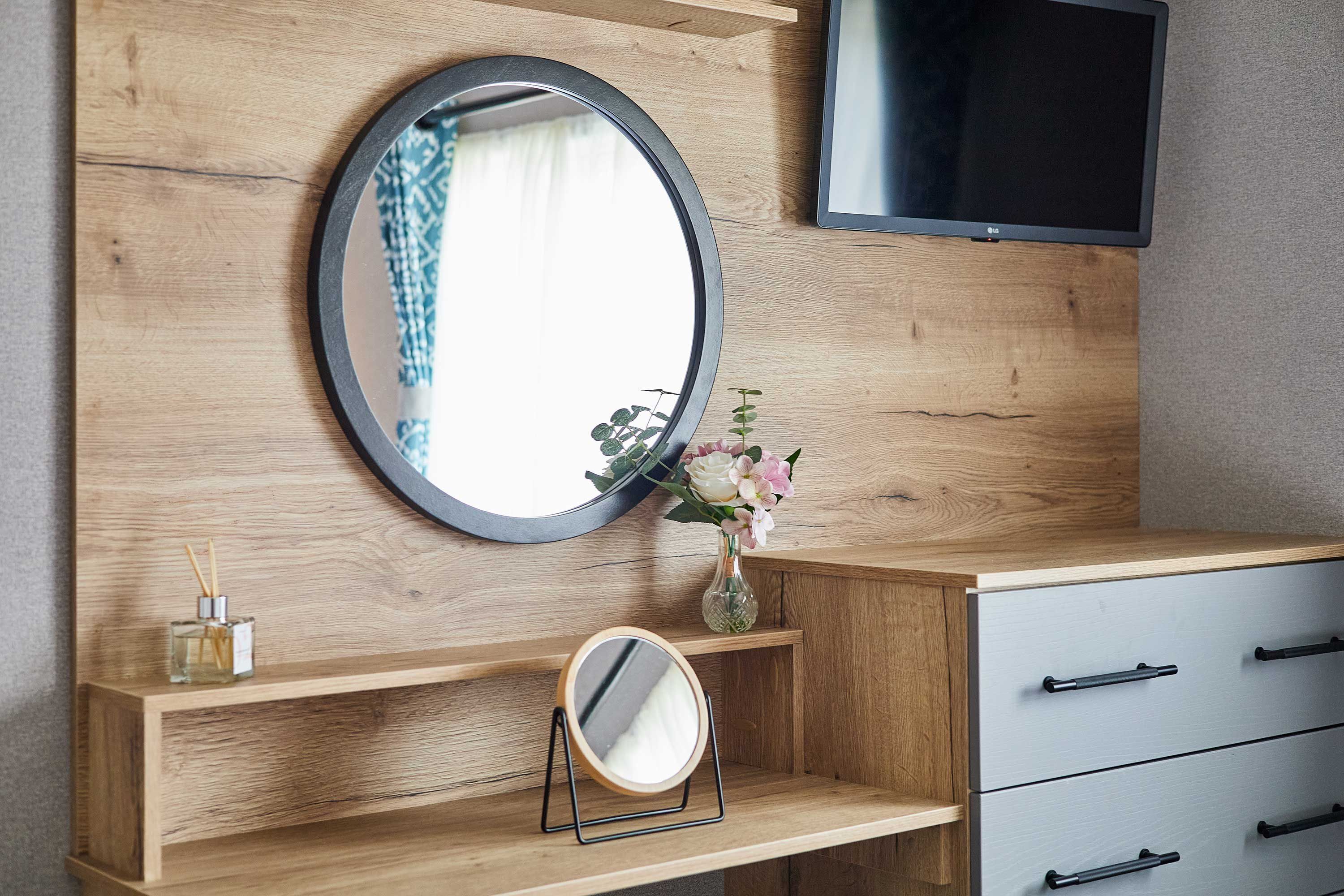 A wooden vanity with a round mirror, a small shelf with a vase of flowers, and a television mounted above.