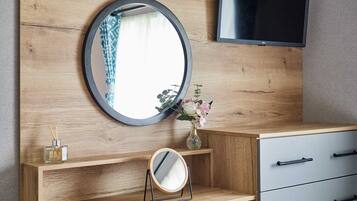 A wooden vanity with a round mirror, a small shelf with a vase of flowers, and a television mounted above.