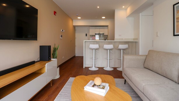 A modern living room with a flat-screen TV, a sofa, a coffee table with magazines, and a kitchen in the background. - w Enchanting 1BR w Balcony in Prime Miraflores (Lima)