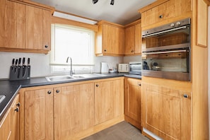 A compact kitchen with wooden cabinets, a built-in oven, and a window with blinds. - Host Stay Plane View (Morpeth)