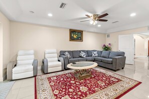 Ein Wohnzimmer mit einem grauen Sofa, weißen Sesseln, einem Glastisch, einem Teppich mit roten und weißen Mustern, einem Deckenventilator und einem Bild an der Wand.