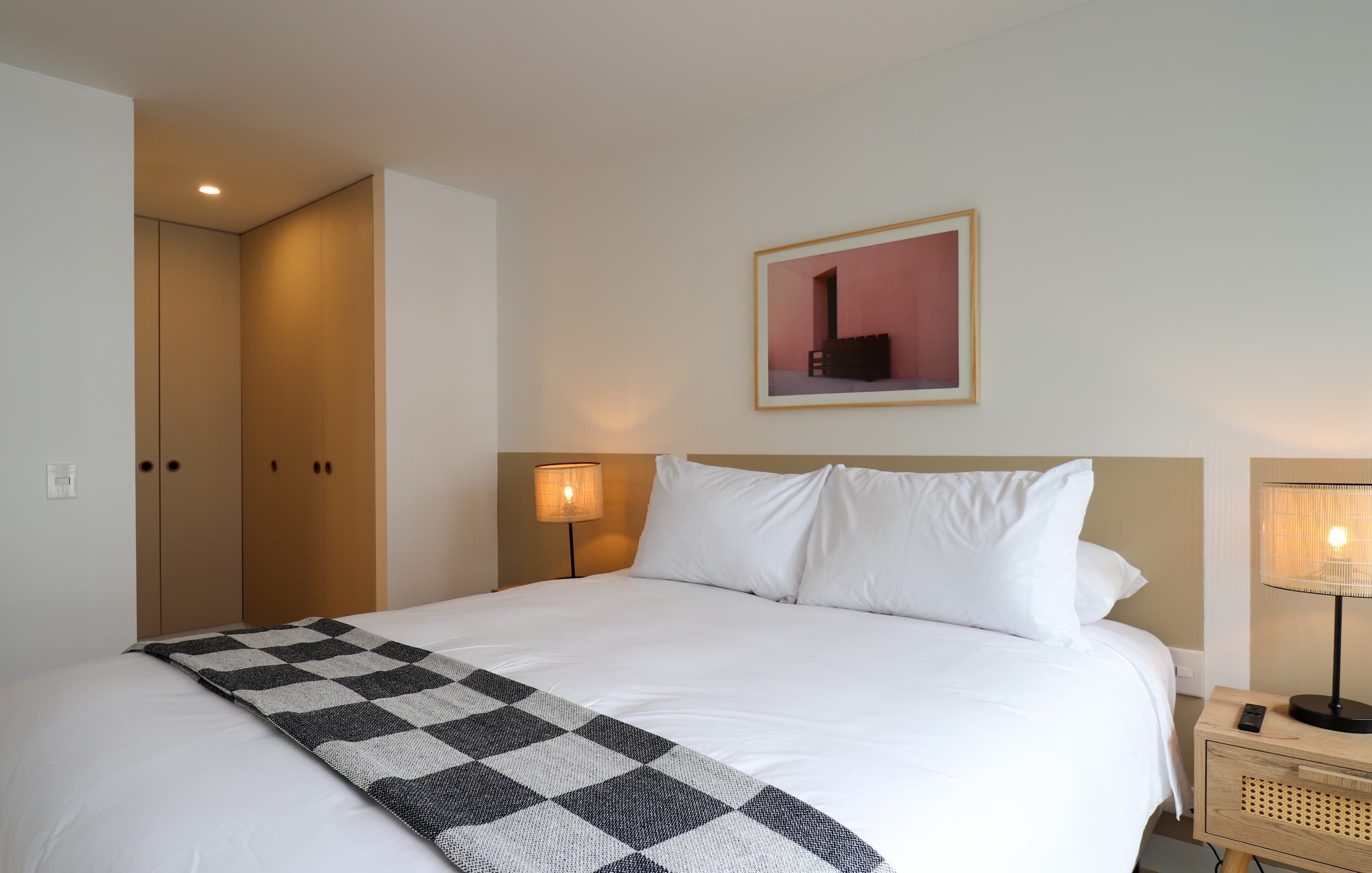 A hotel room with a large bed, a checkered throw, a bedside lamp, a framed picture on the wall, and a wardrobe.
