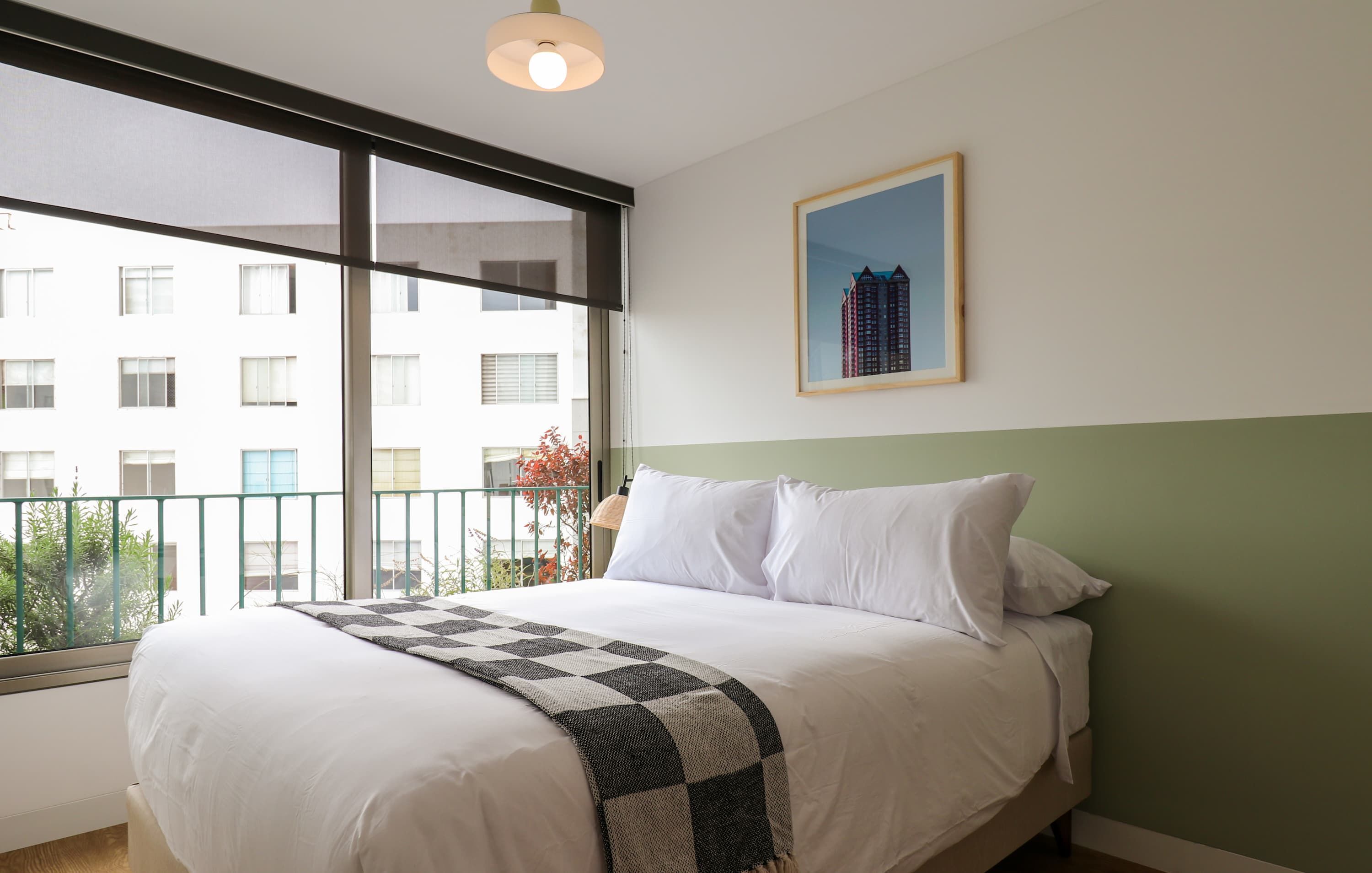 A hotel room with a large window, a bed with white linens, a framed picture on the wall, and a view of a building outside.