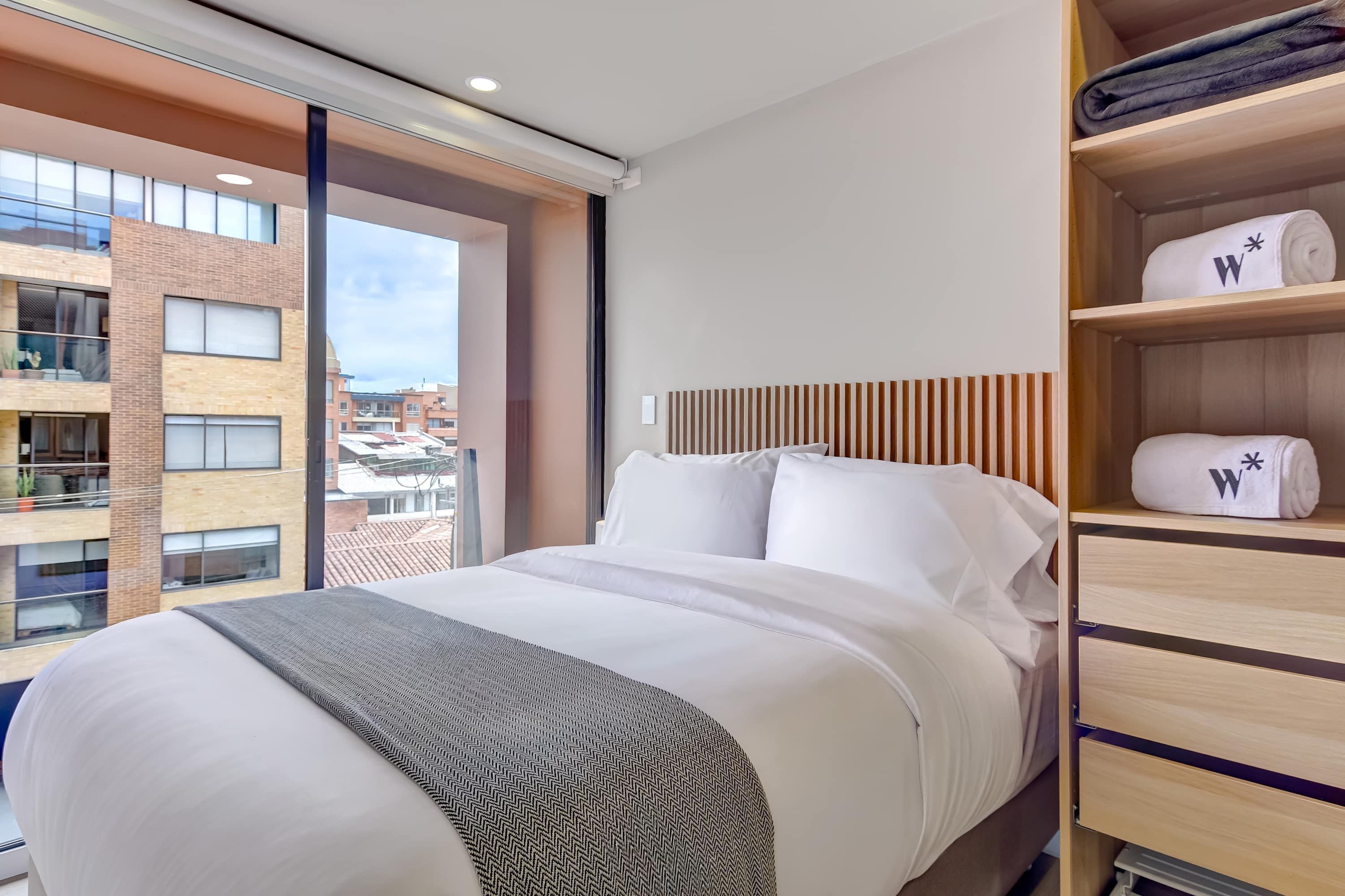 A hotel room with a large bed, a wooden headboard, a window with a view of buildings, and a shelf with towels.