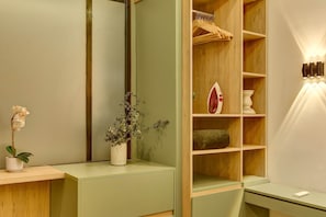 Quarto com espelho, pequena mesa com planta em vaso, estante de madeira com roupas penduradas e luminária fixada na parede.
