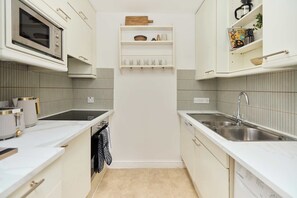 A compact kitchen with white cabinets, a microwave, a sink, and a small shelf with kitchenware. - The Two-Bedroom Edit (Greater London)