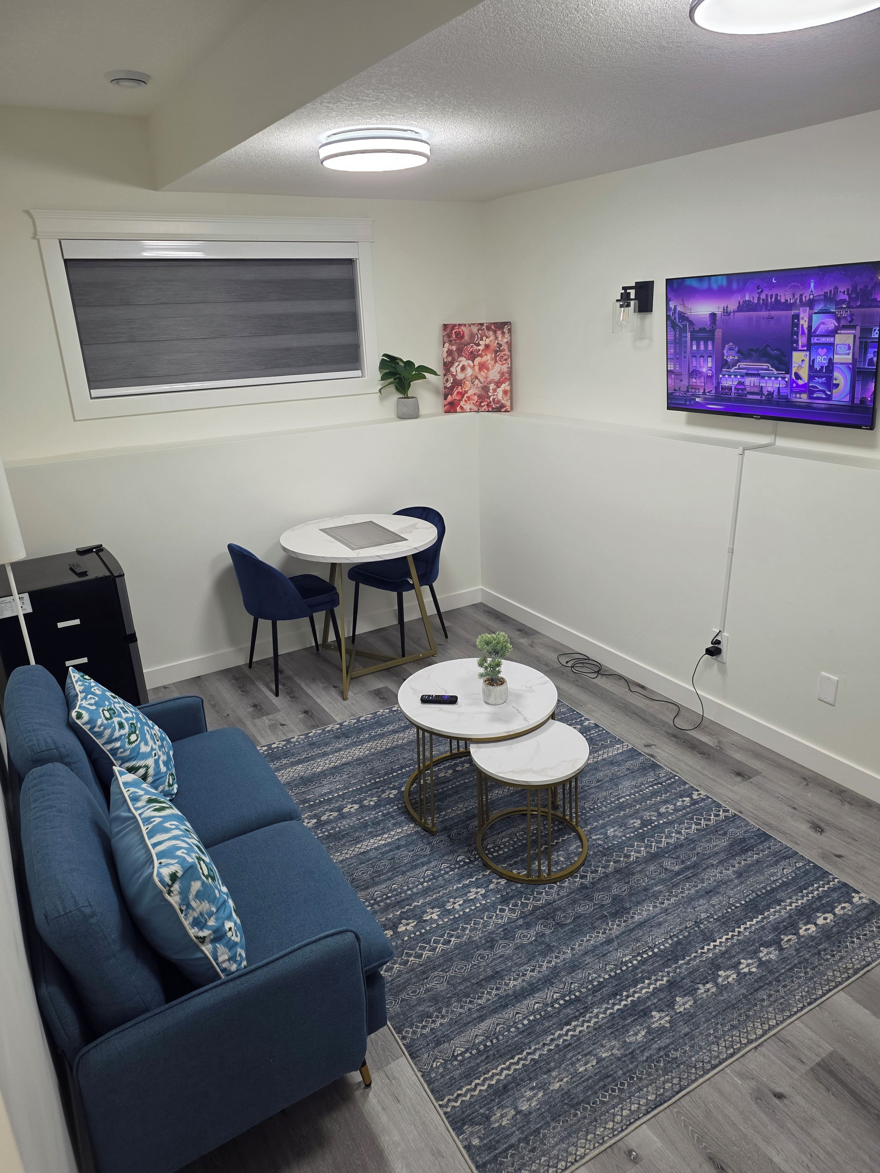 A modern living room with a blue sofa, a small dining table with chairs, a TV mounted on the wall, and a window with a view.