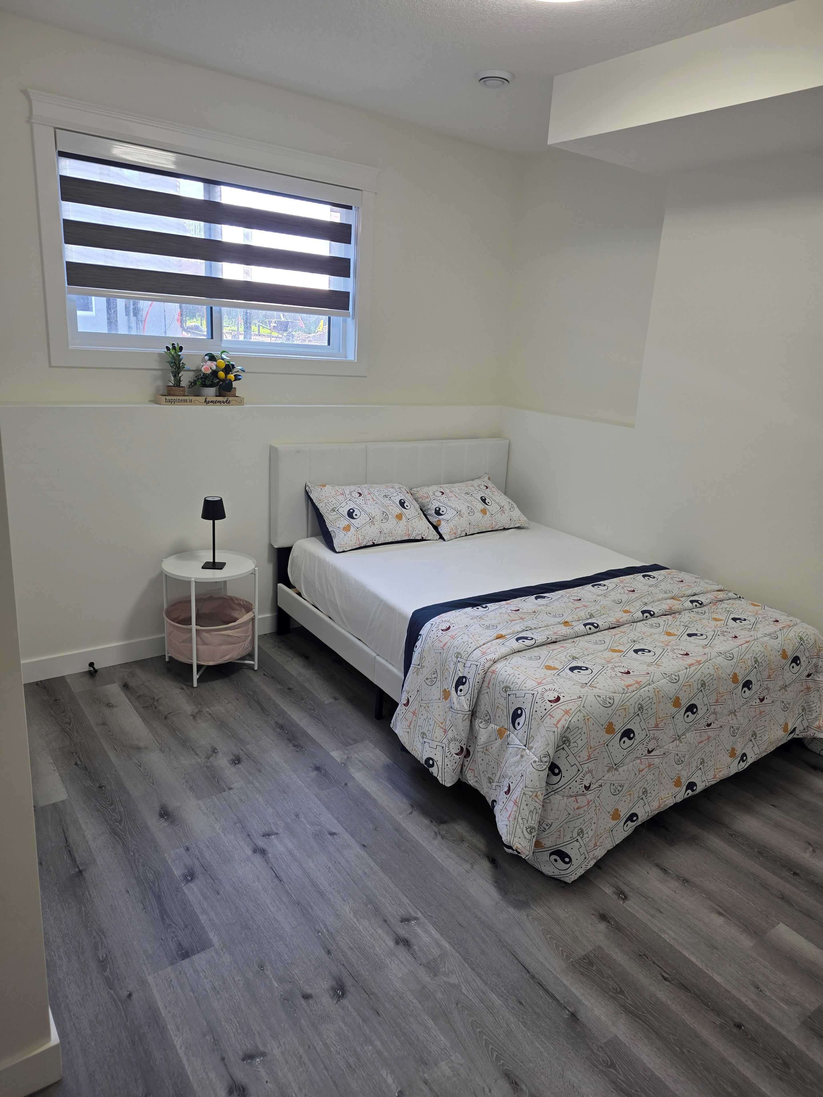 A bedroom with a bed, a bedside table, a lamp, a window with blinds, and a small plant on the windowsill.