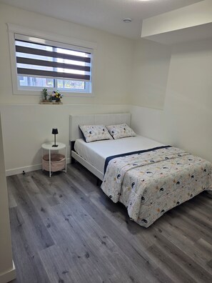 A bedroom with a bed, a bedside table, a lamp, a window with blinds, and a small plant on the windowsill. - Marilyn’s Luxurious Basement Apartment (Saskatoon)