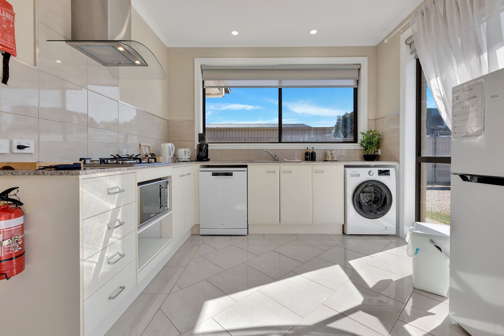 A modern kitchen with white cabinetry, a built-in oven, a dishwasher, a washing machine, and a window with a view of the outside.