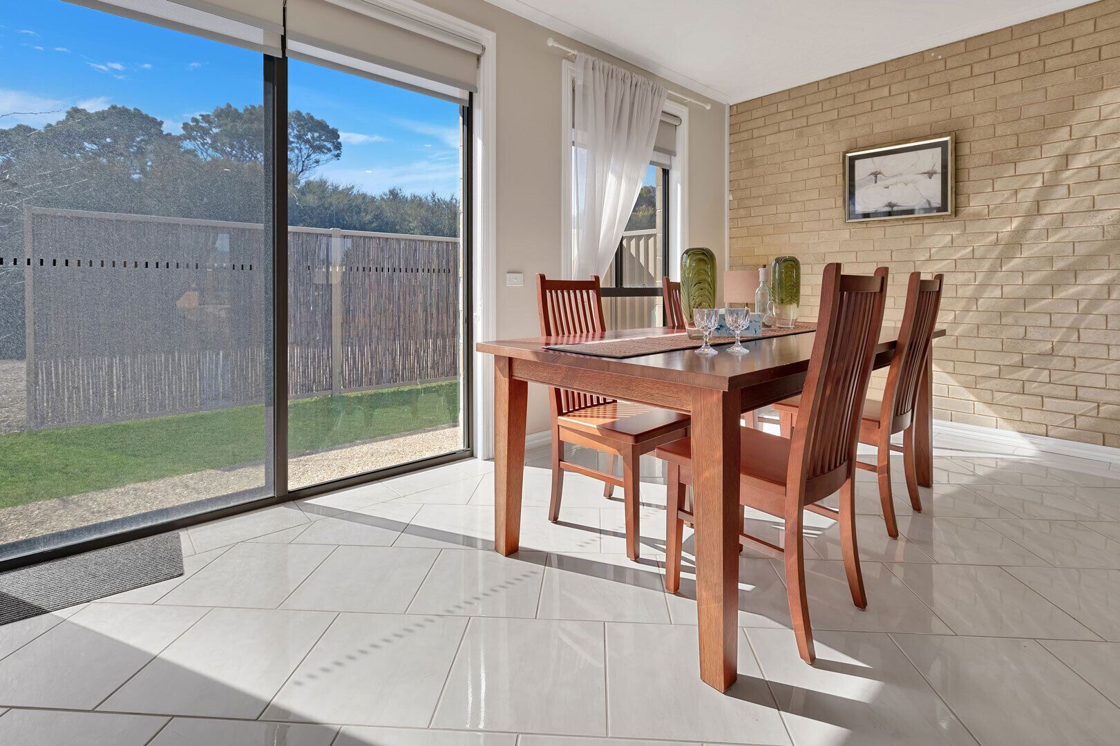 A dining area with a large table and chairs, a view through a sliding glass door, and a brick wall with a framed picture.
