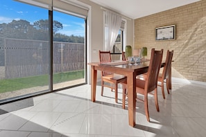 Un coin repas avec une grande table et des chaises, une vue à travers une porte-fenêtre coulissante et un mur de briques orné d’un tableau encadré.